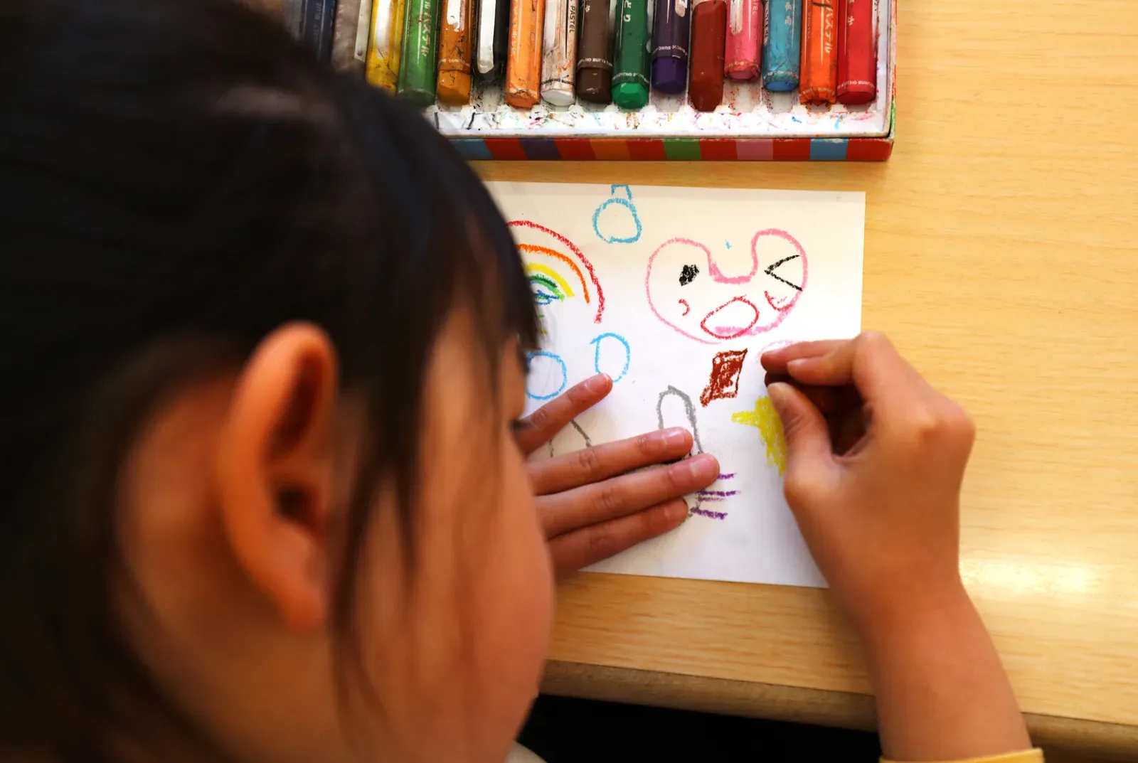 オイルパステルを握って夢中で絵を描く子どもの手元と、使い込んだクレヨンのケース